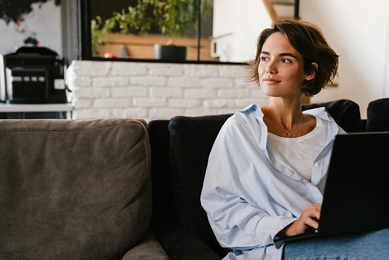 Beautiful young smiling woman in casual wear working on laptop computer while sitting on couch in modern workspace