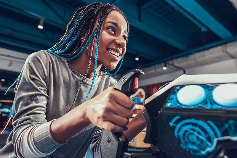 African American girl piloting blue plane in arcade. Beautiful girl enthusiastically plays a video game with a joystick in an entertainment center.